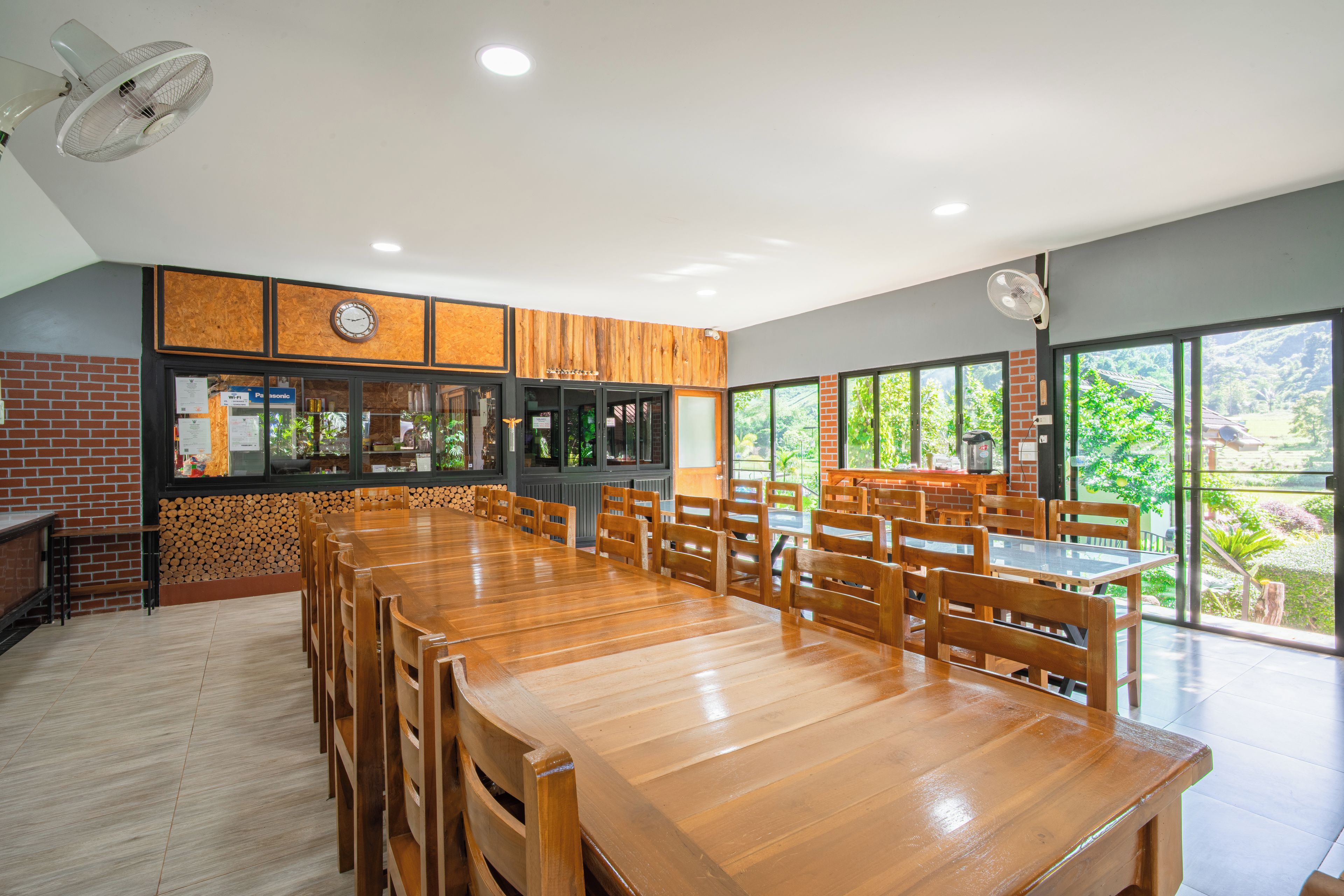 Un comedor amplio con una larga mesa de madera y sillas, pared de ladrillo y ventanas grandes que ofrecen vistas al verdor.