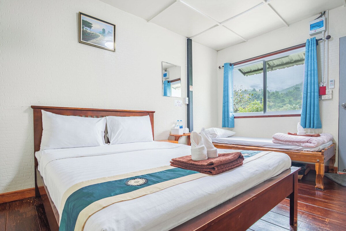 A hotel room with a large bed, a smaller bed, a wooden bench, a window with curtains, and a view of greenery outside.