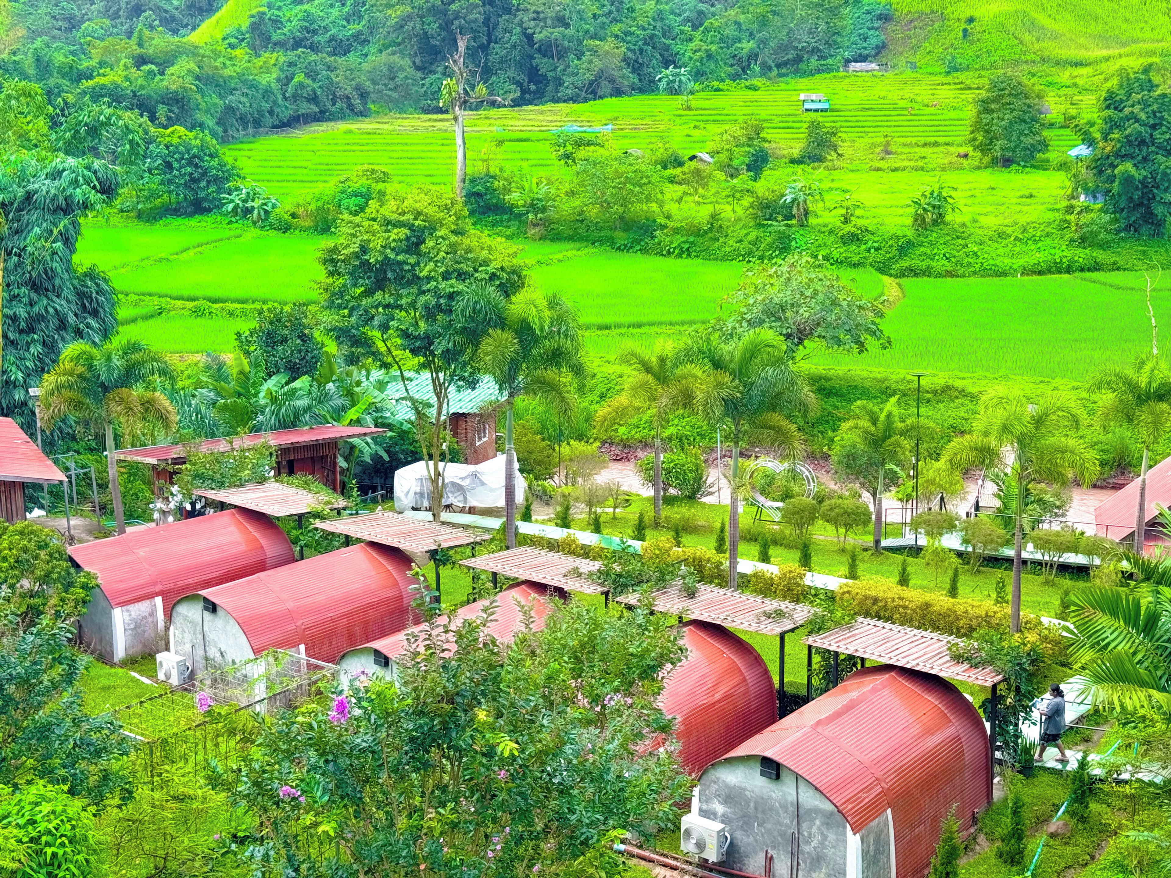 Paisaje rural con campos verdes, algunas edificaciones con techos rojos y una persona caminando.