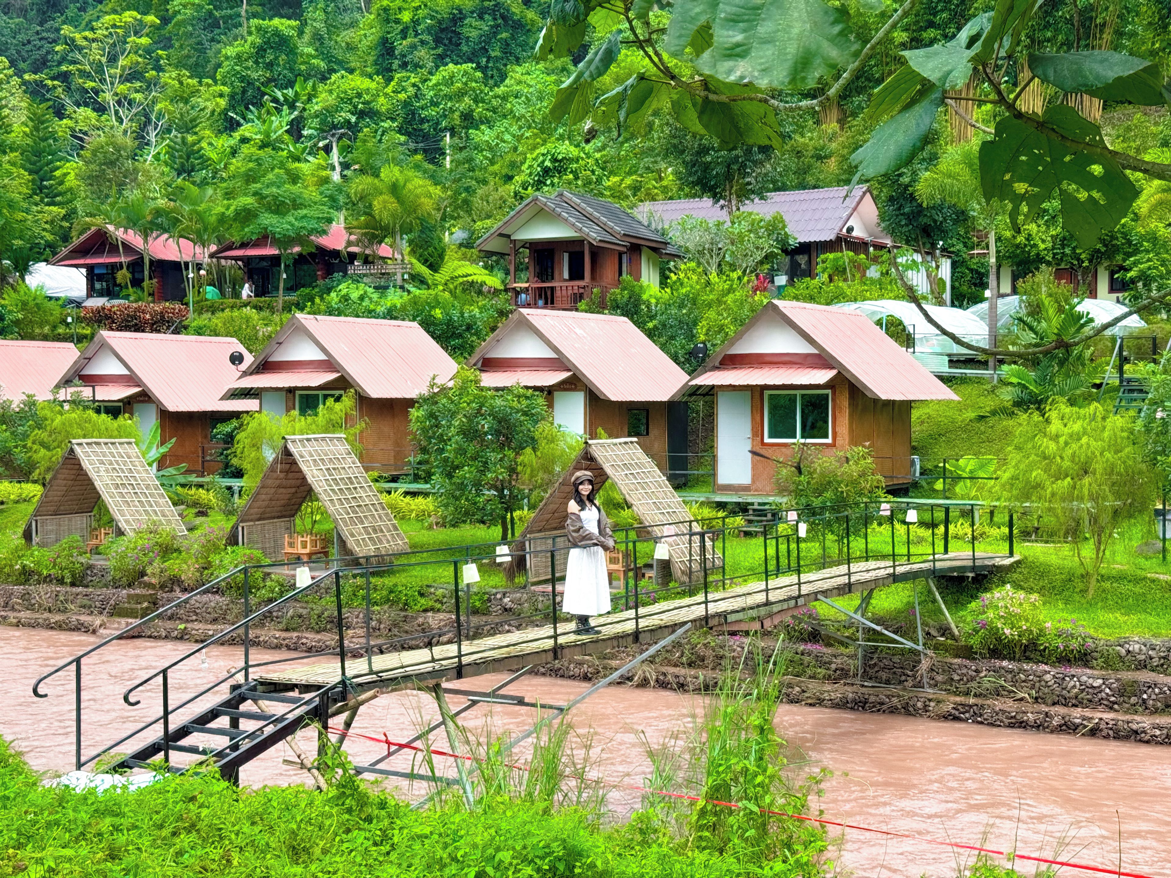 A series of wooden cabins with pitched roofs, surrounded by lush greenery and a river.