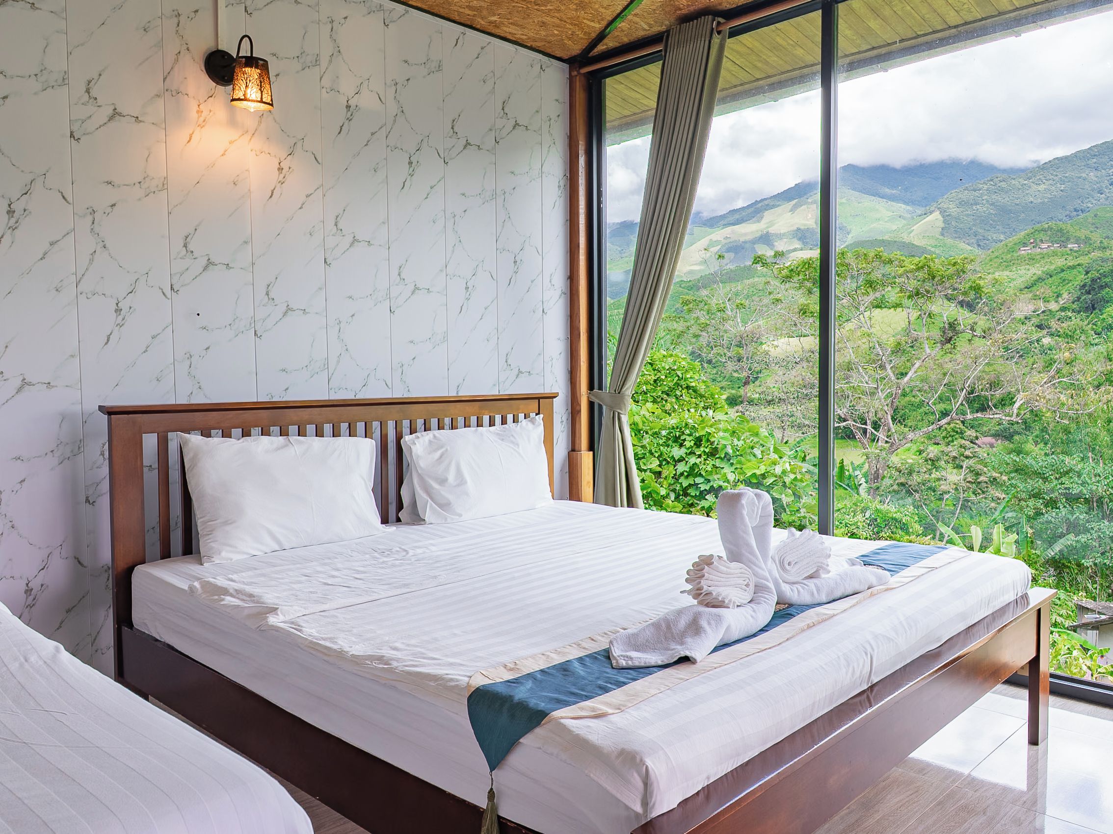 A bedroom with a large bed, a nightstand, a wall-mounted lamp, and a view of a mountainous landscape.