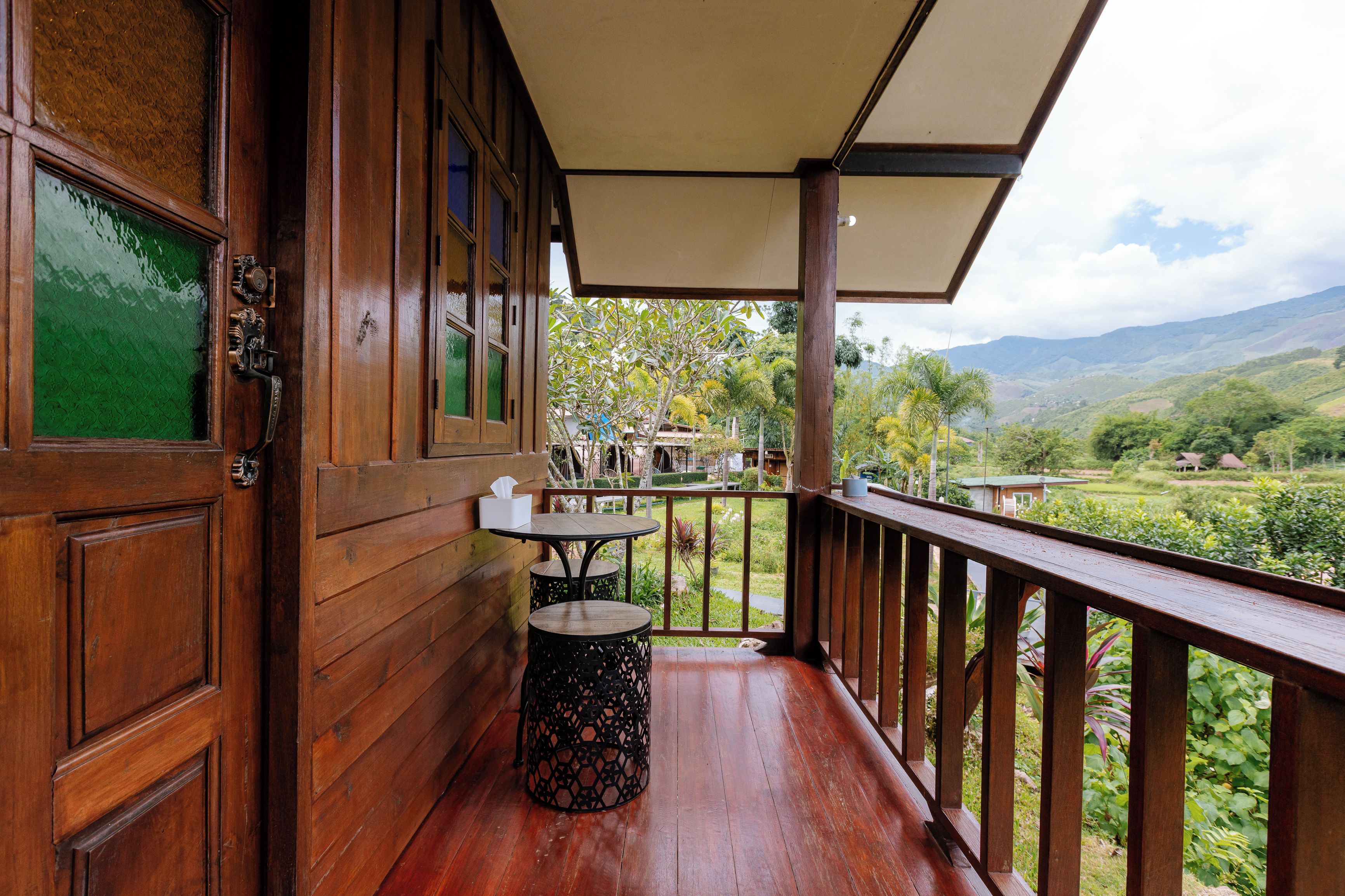 A wooden balcony with a table and chairs, a trash bin, and a view of a lush landscape.