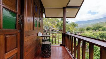 A wooden balcony with a table and chairs, a trash bin, and a view of a lush landscape.
