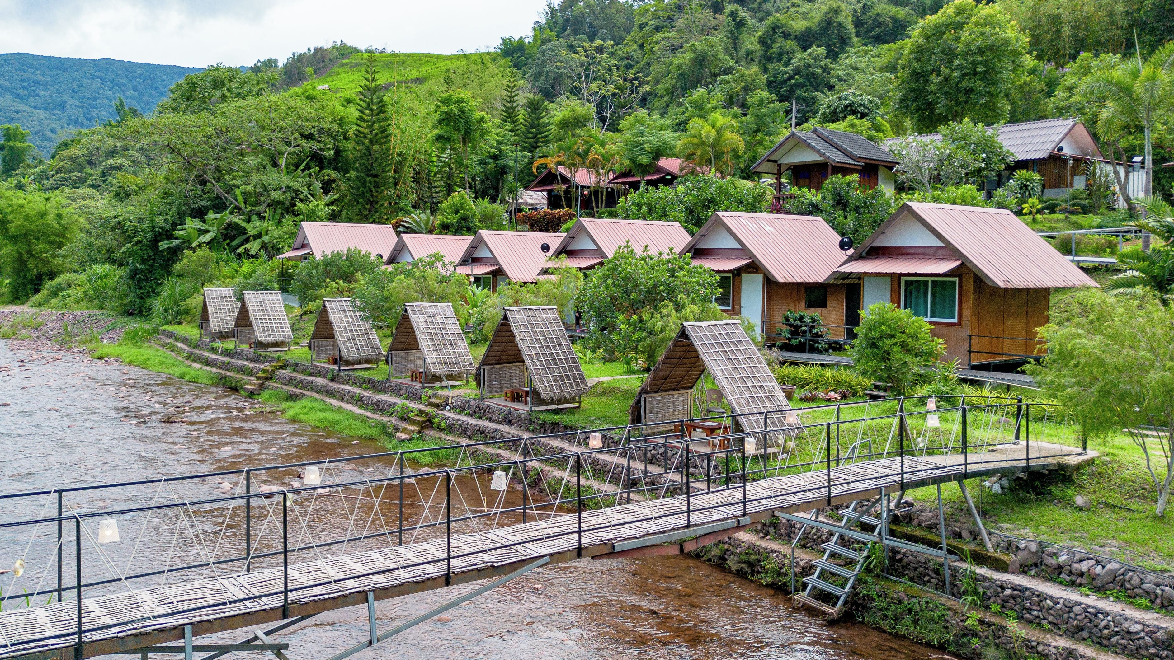A series of traditional-style cottages with sloped roofs, a wooden bridge, and a river in the foreground.