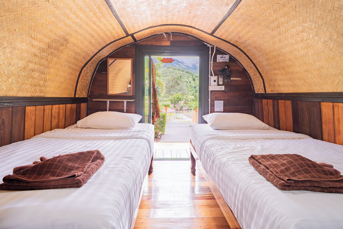 A room with two beds, a wooden ceiling, and a view of greenery outside.