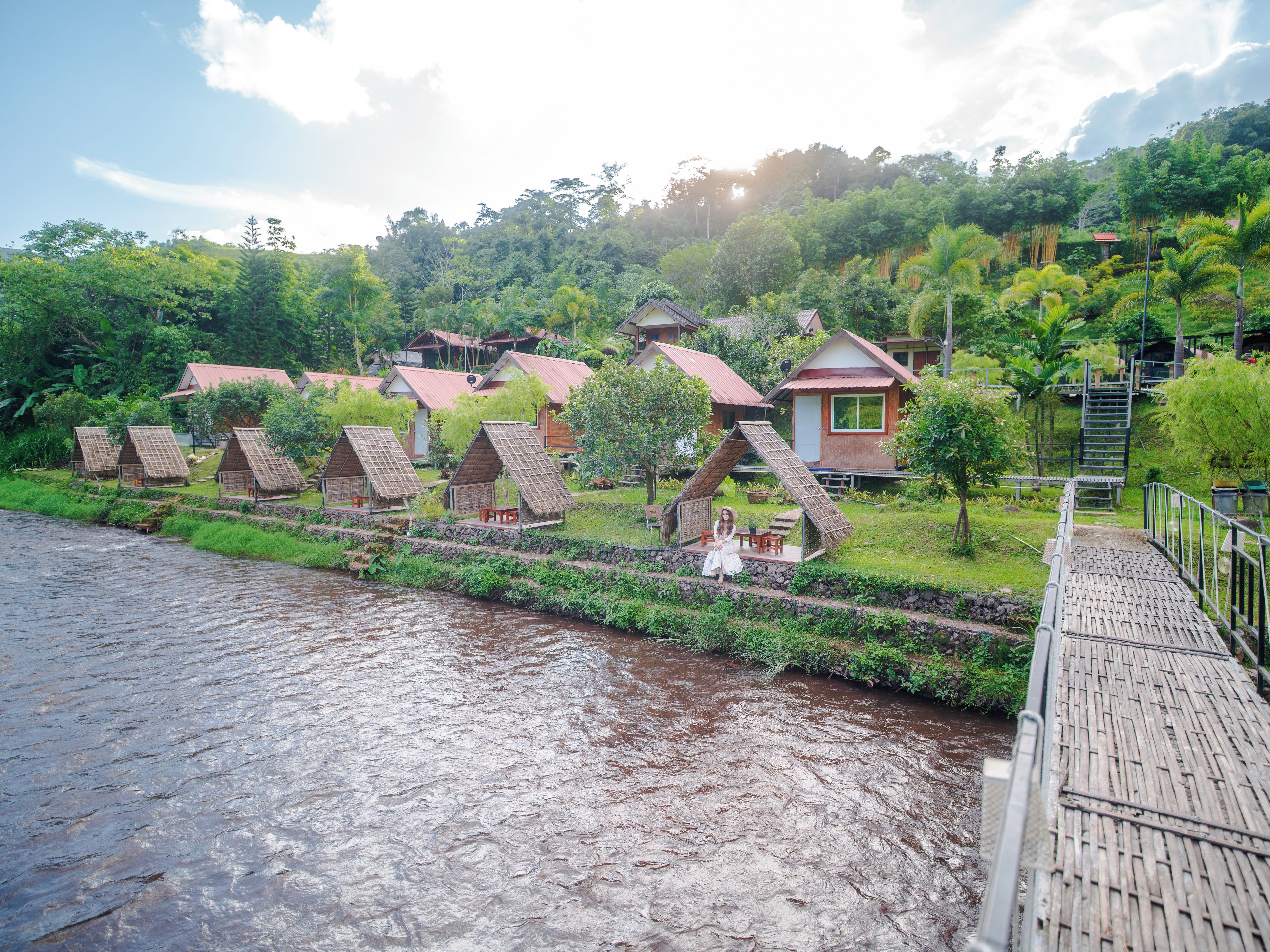 Una hilera de cabañas ecológicas de estilo A, con un río al frente, un puente de madera y abundante vegetación en el fondo.