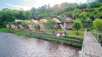 Una hilera de cabañas ecológicas de estilo A, con un río al frente, un puente de madera y abundante vegetación en el fondo.