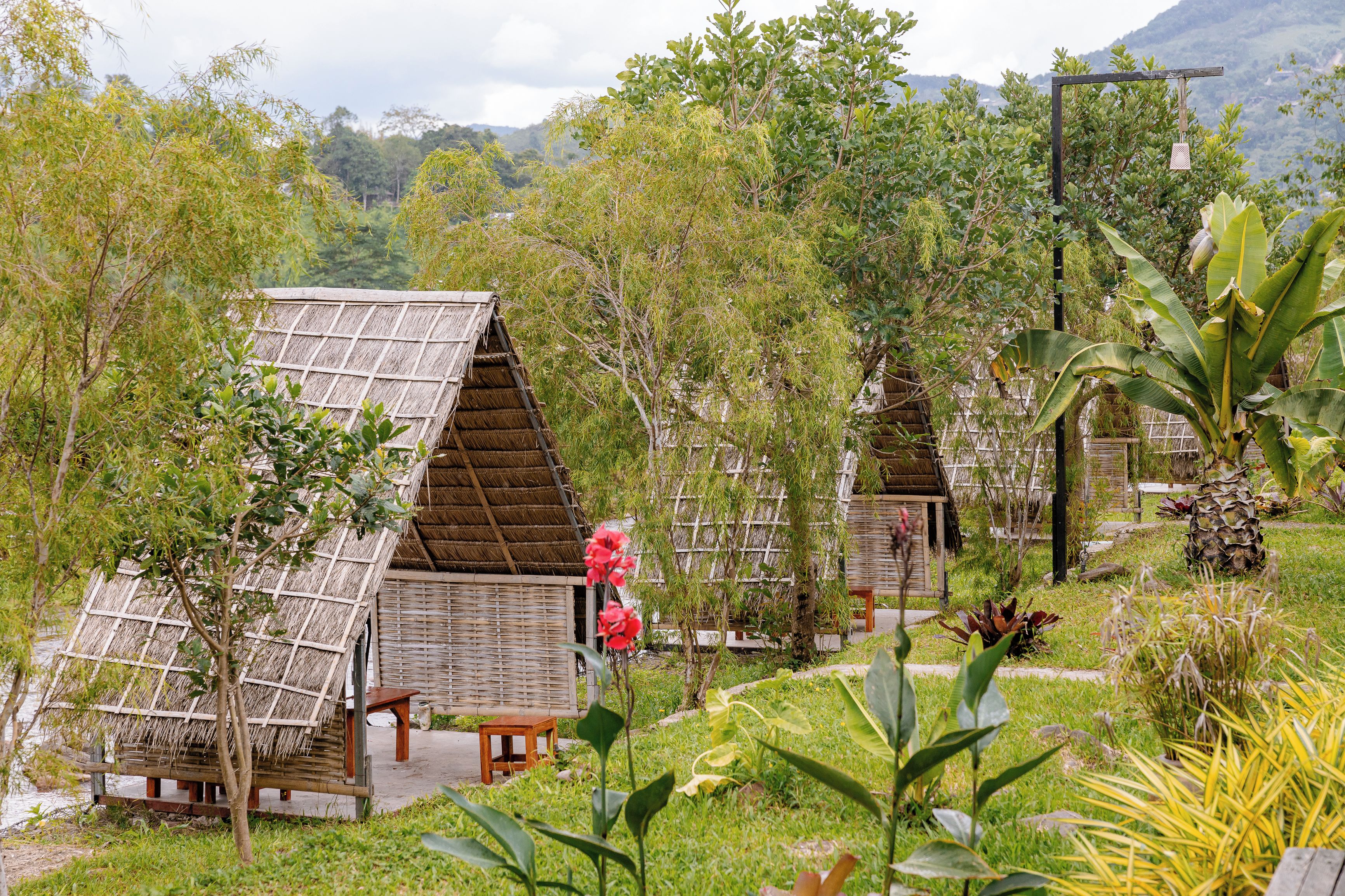 A rustic outdoor setting with thatched-roof huts surrounded by lush greenery and a mountainous backdrop.
