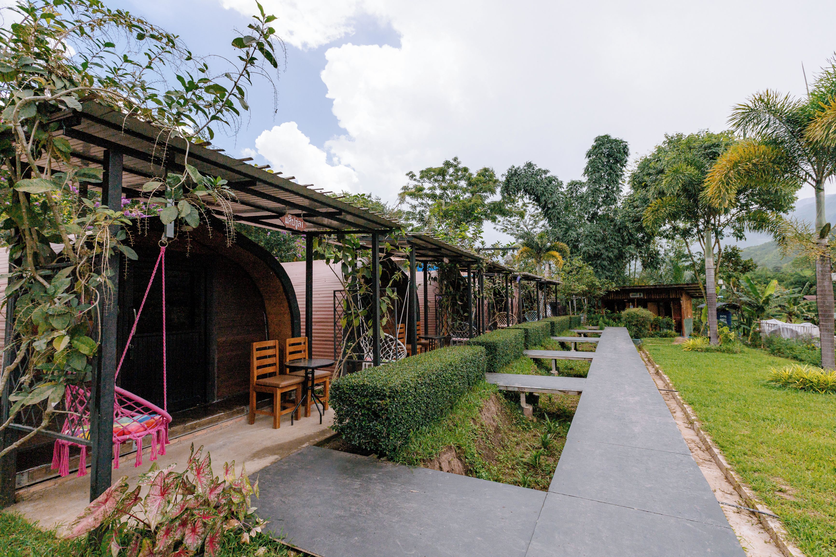 A covered outdoor seating area with tables and chairs, surrounded by greenery and a pathway.