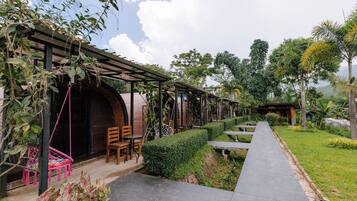 A covered outdoor seating area with tables and chairs, surrounded by greenery and a pathway.