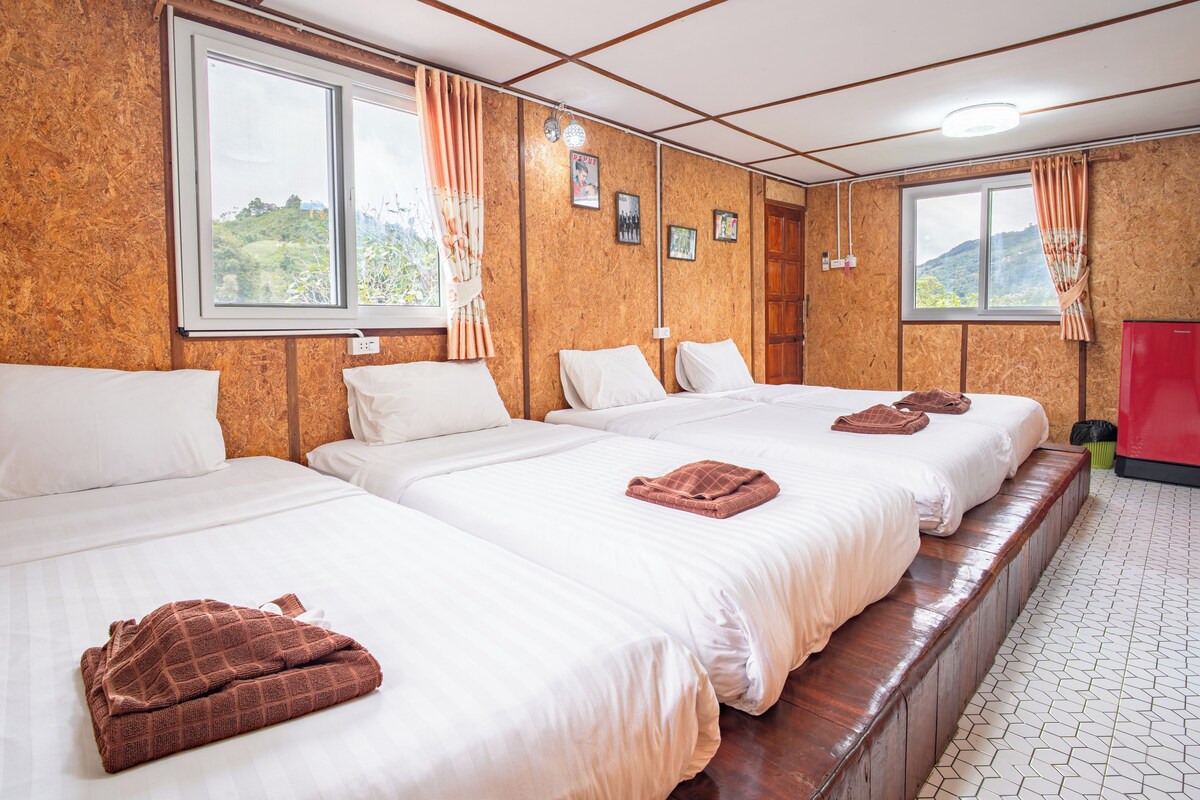 A hotel room with three beds, wooden paneling, a red refrigerator, and a view of greenery outside.