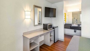 Habitación de hotel compacta con piso de madera, kitchenette con microondas, televisor de pantalla plana y baño con lavamanos y espejo.