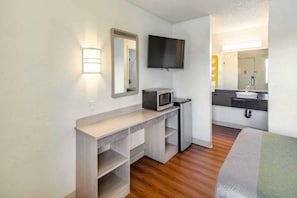A compact hotel room with a wooden floor, a kitchenette with a microwave, a flat-screen TV, and a bathroom with a sink and mirror. - 1 Queen Bed | Non-Smoking (Clarkston)