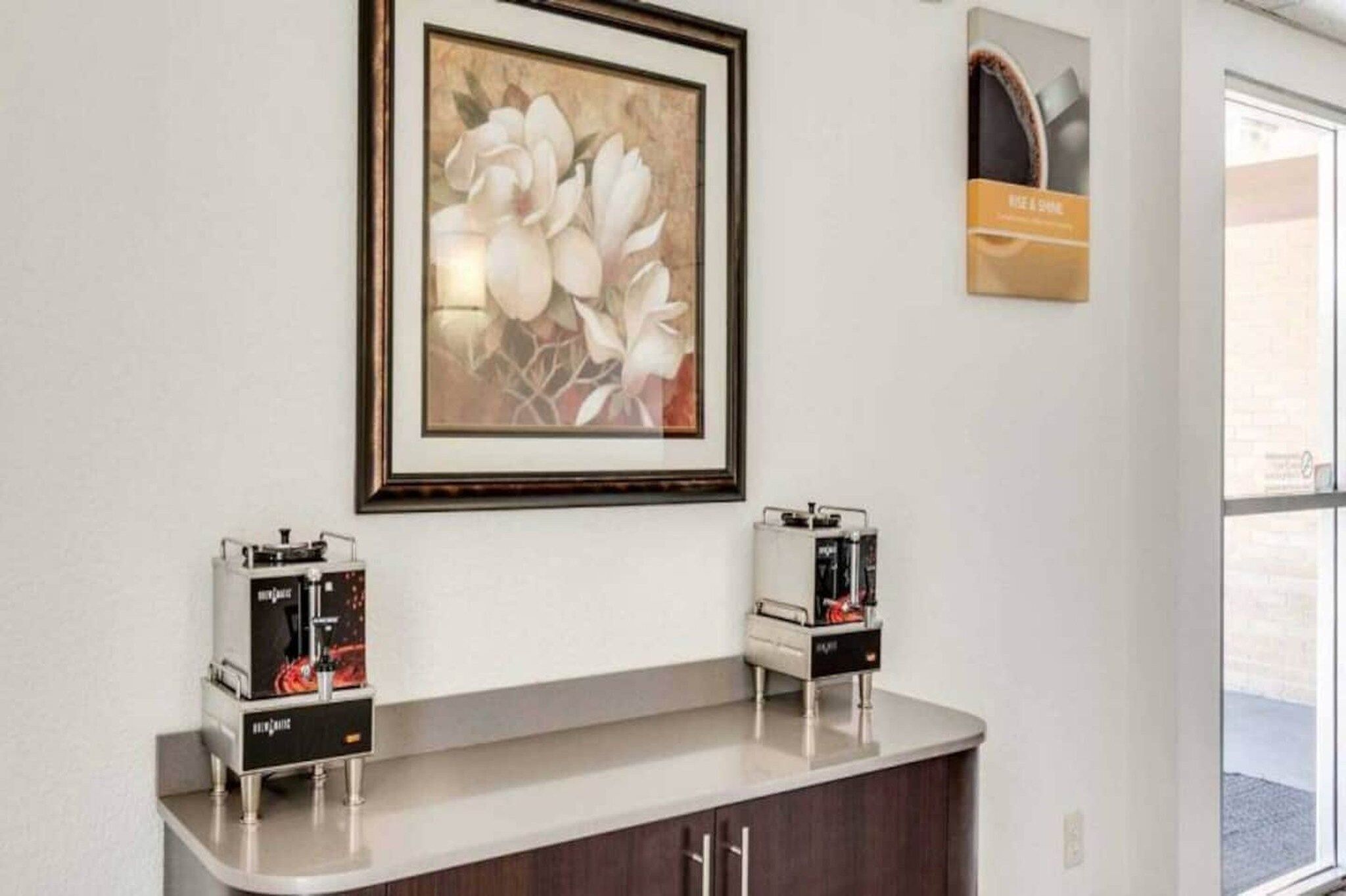 Un área de recepción con una obra de arte floral enmarcada sobre un mostrador, dos máquinas de café y un cartel con el texto 'RISE & SHINE'.