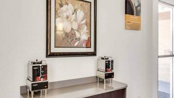 Un área de recepción con una obra de arte floral enmarcada sobre un mostrador, dos máquinas de café y un cartel con el texto 'RISE & SHINE'.