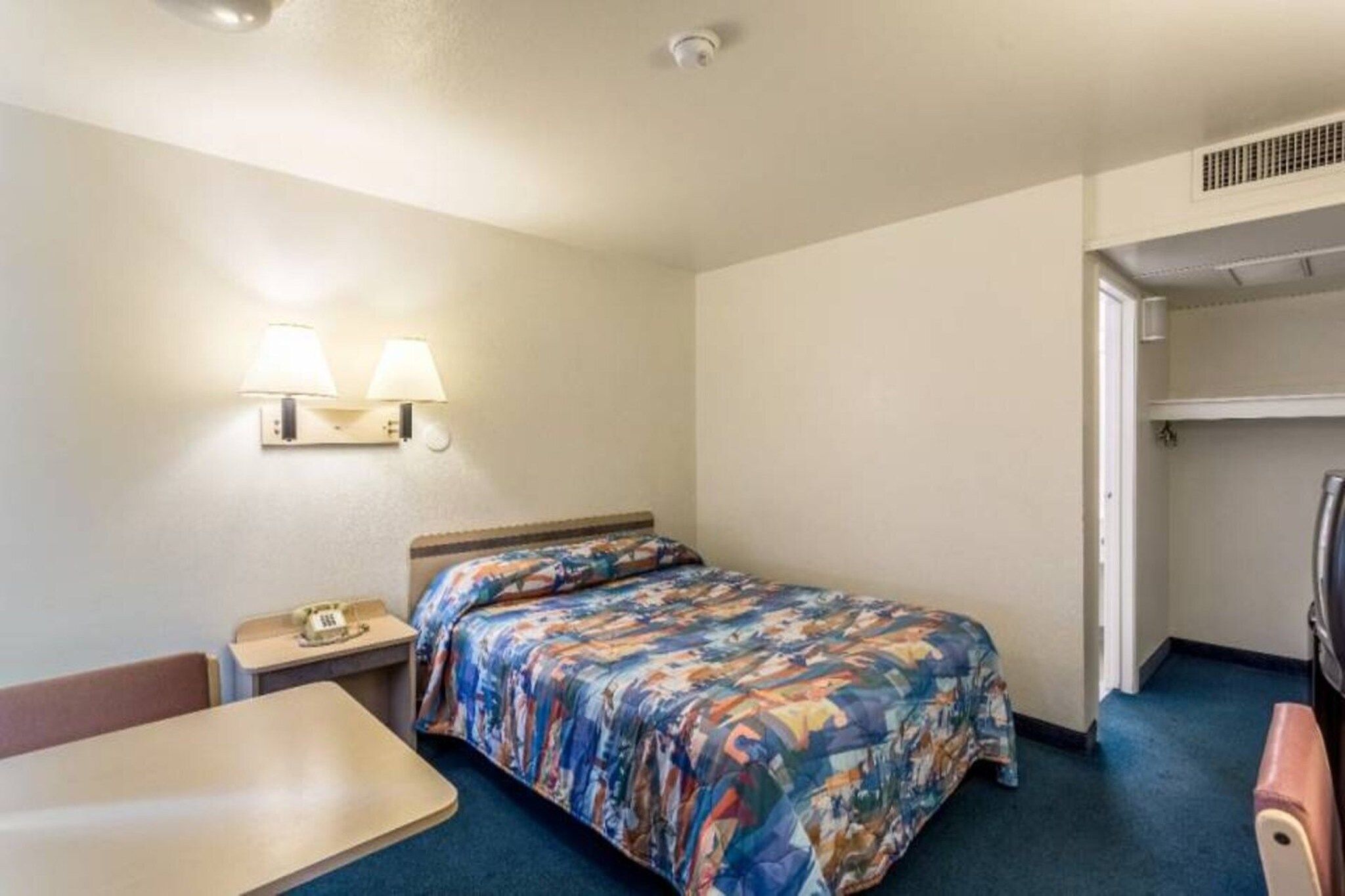 Quarto de hotel com cama, escrivaninha, cadeira, mesa de cabeceira e luminária na parede.