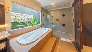 A modern bathroom with a freestanding bathtub, a glass shower enclosure, and a window with a view of greenery. - Cozzy Dewa Villa Sunset Road (Badung)