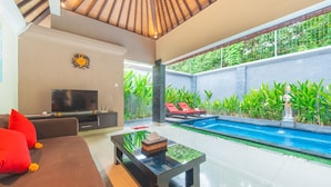 A modern living area with a TV, sofa, and coffee table, a pool with red lounge chairs, and a view of lush greenery. - Cozzy Dewa Villa Sunset Road (Badung)