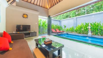 Un salón moderno con televisor, sofá y mesa de centro, una piscina con sillas reclinables rojas y exuberante vegetación.