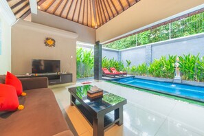Ein modernes Wohnzimmer mit Fernseher, Sofa und Couchtisch, ein Pool mit roten Liegestühlen und Blick auf üppiges Grün.