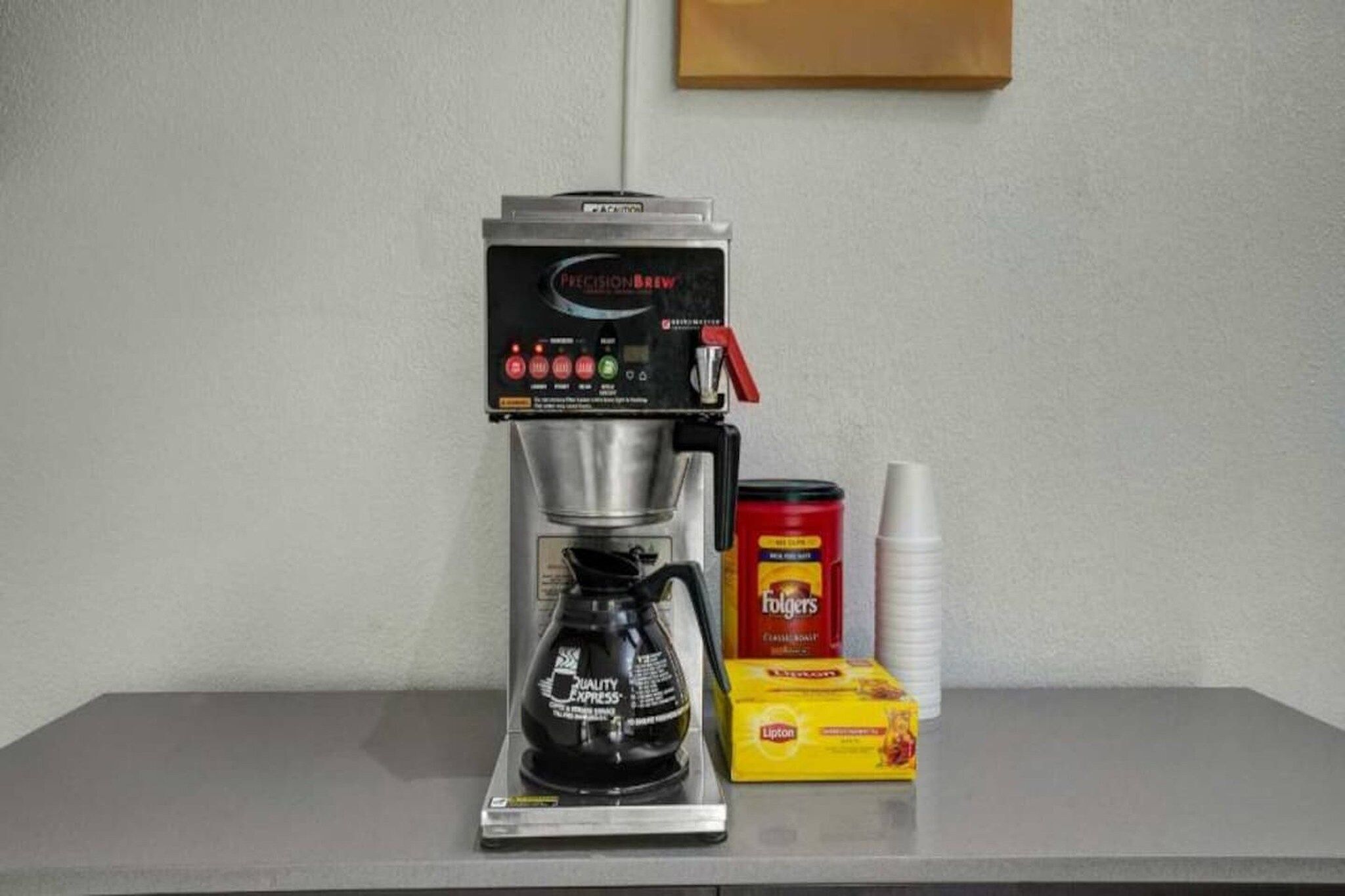 Een koffieapparaat met een koffiekan, een doos Folgers koffie, een blik Lipton thee en een stapel witte papieren bekers op een tafel.