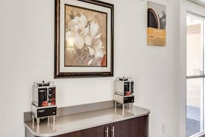 A reception area with a framed floral artwork above a counter, two coffee machines, and a sign with the text 'RISE & SHINE'.