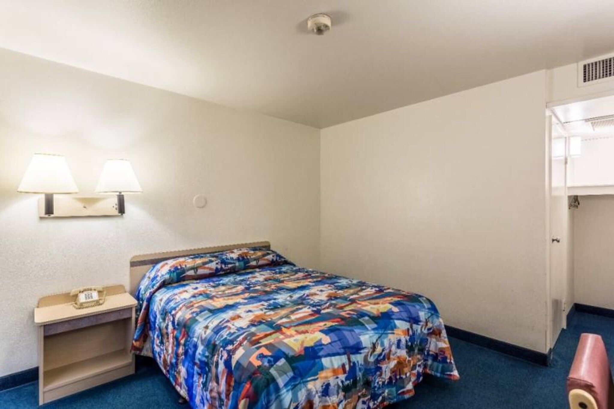 A single bed with a colorful bedspread, a nightstand with a lamp, and a door to a closet.
