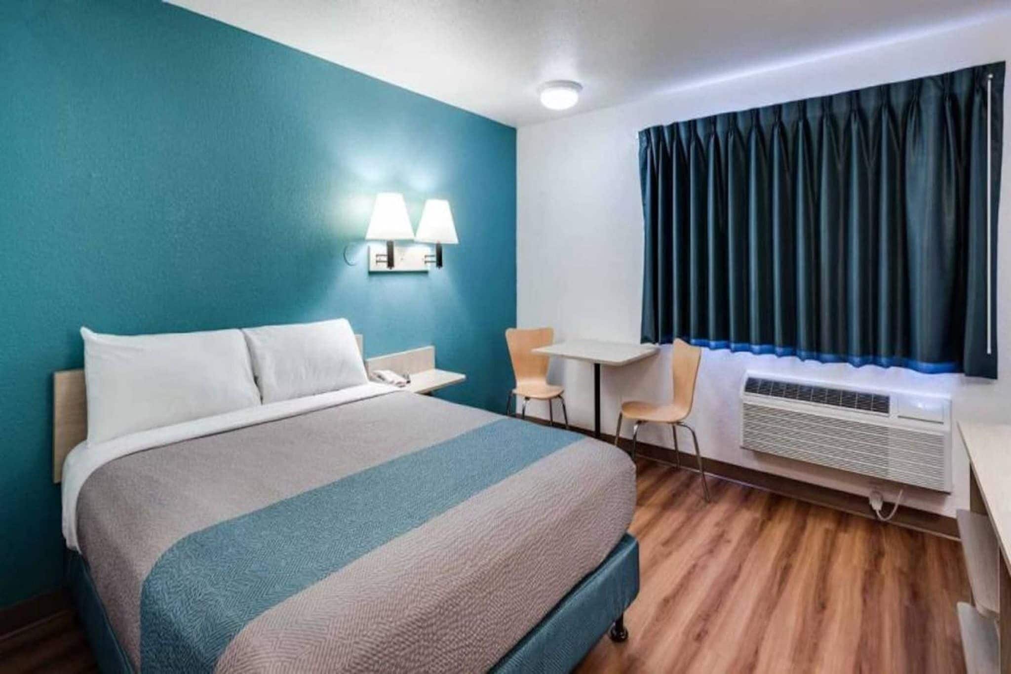 A hotel room with a bed, desk, chairs, and an air conditioning unit.