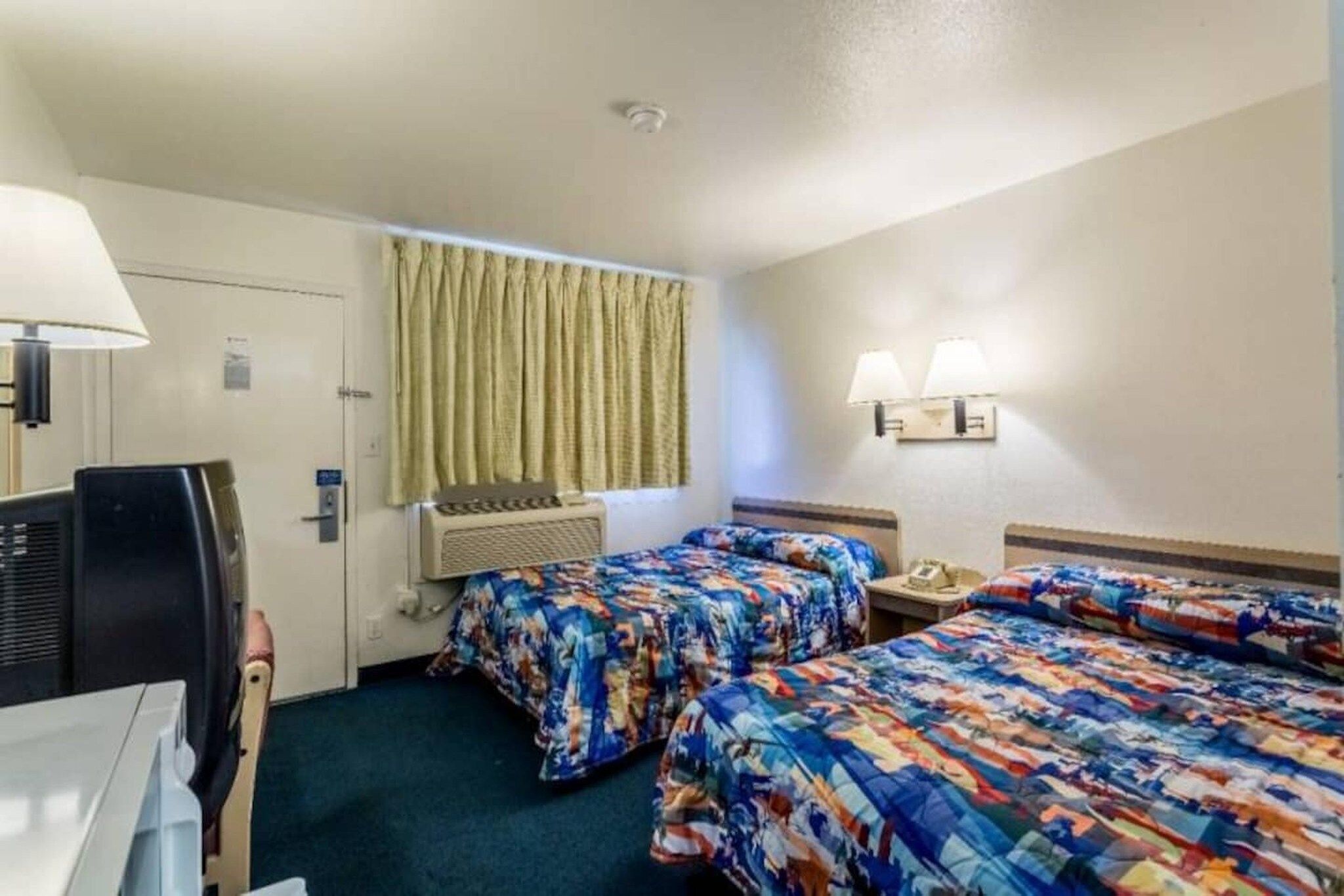 A hotel room with two beds, a desk, a chair, a television, an air conditioning unit, and a window with curtains.