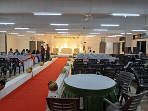 A conference room with rows of chairs, a stage with a red carpet, and a ceiling with linear lighting fixtures.