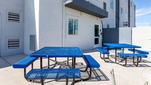 An outdoor area with multiple blue picnic tables and benches, a building with windows and a door, and a clear sky. - Cozy 2-Bed Queen|Free Parking|Wifi (Roswell)