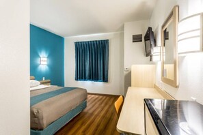 Quarto de hotel com cama, escrivaninha, cadeira, televisão e janela com cortinas.
