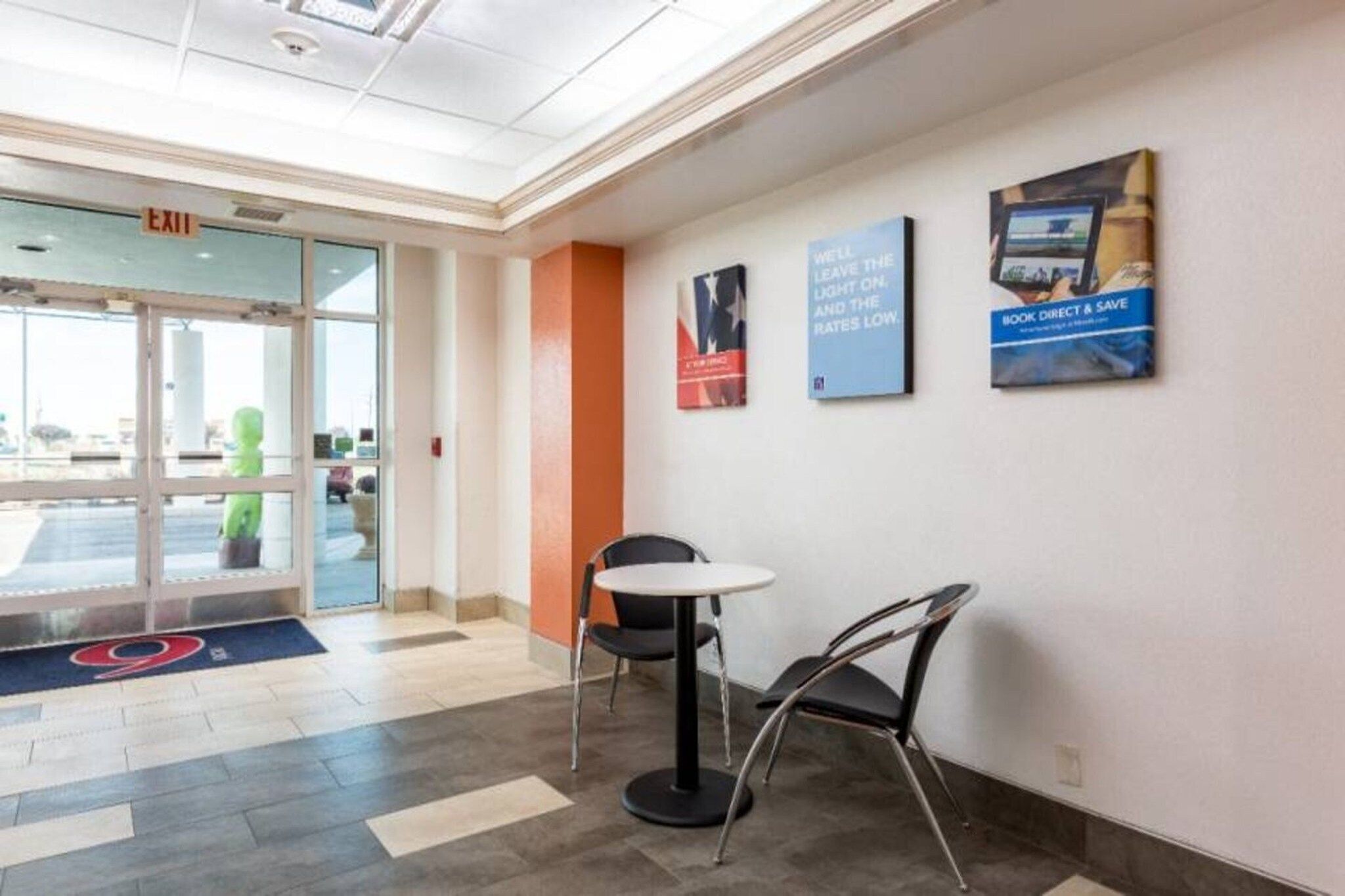 A hotel lobby with a reception desk, a chair, and a table. There is a glass door entrance and a blue exit sign.