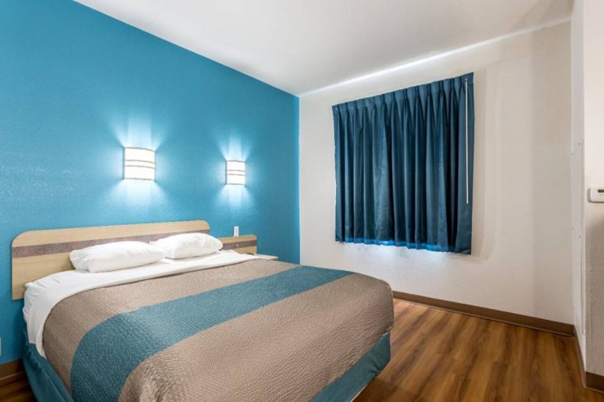 A hotel room with a bed, two wall-mounted lights, a window with curtains, and wooden flooring.
