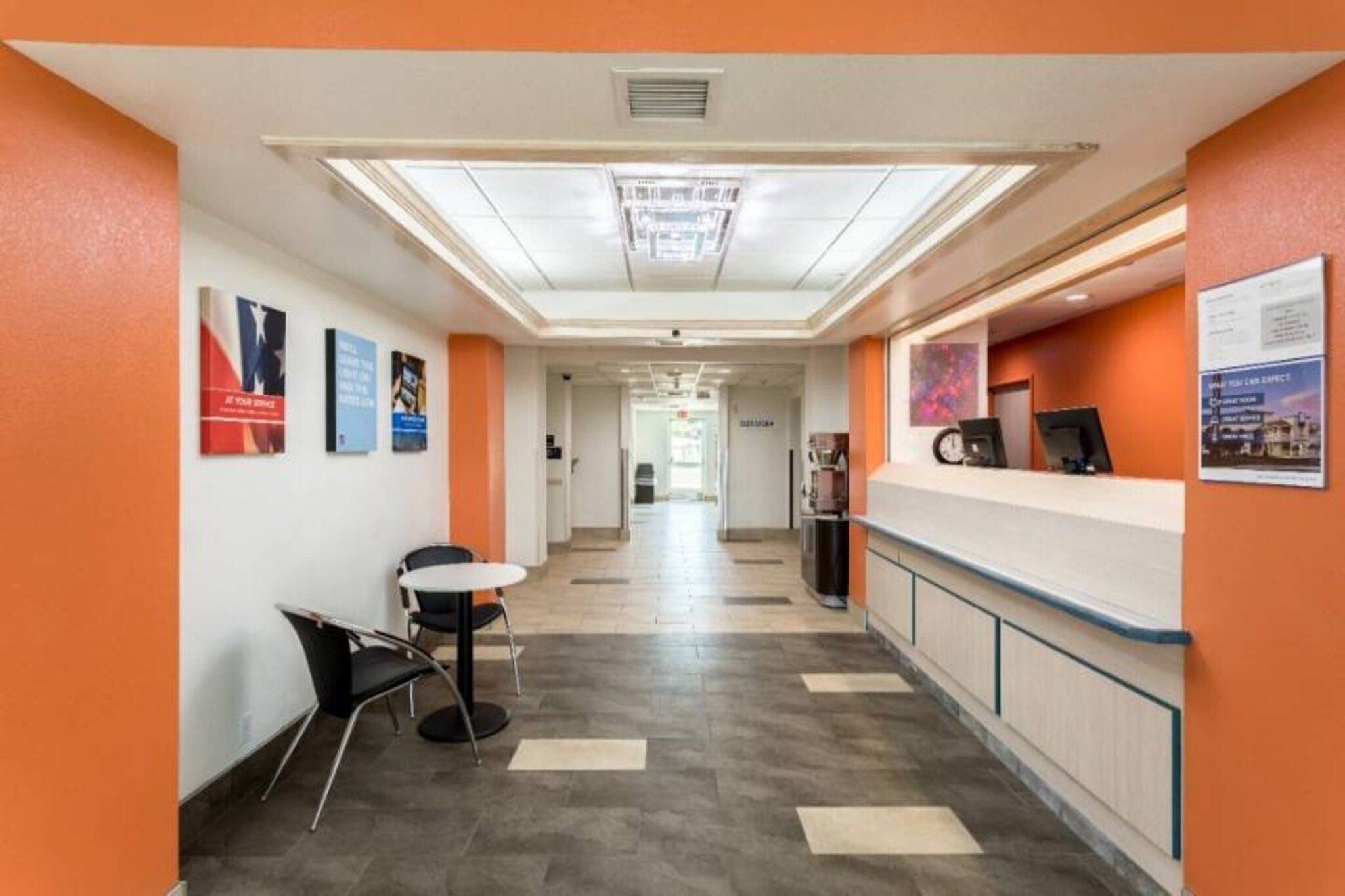 A hotel corridor with a reception desk, chairs, and informational posters on the walls.
