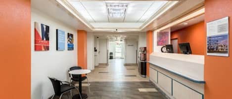 A hotel corridor with a reception desk, chairs, and informational posters on the walls.