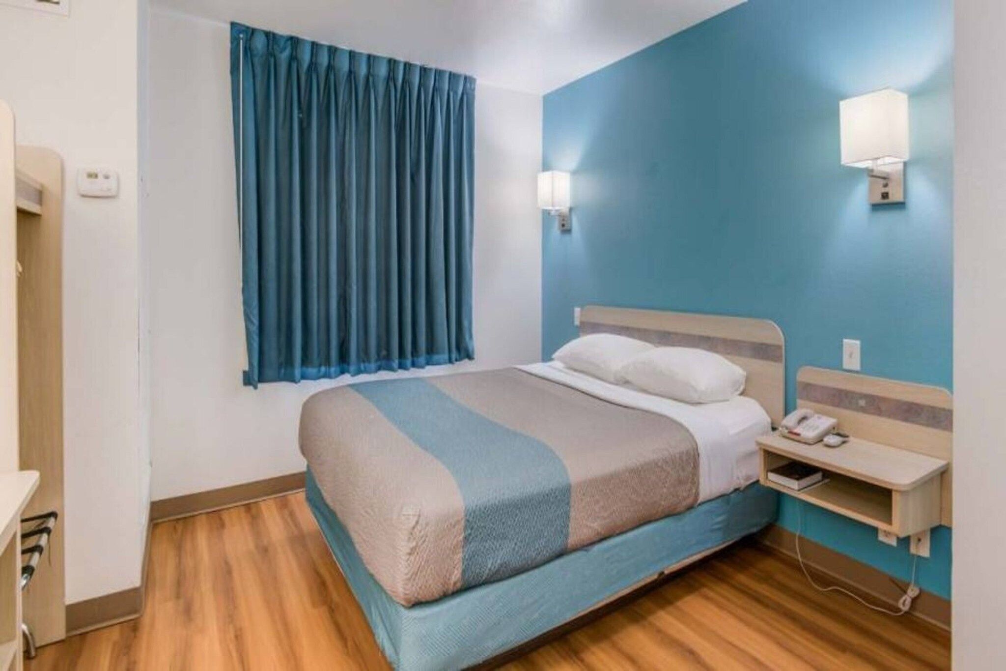 Ein Hotelzimmer mit Bett, Nachttisch, Lampe und einem fenster mit Vorhängen.