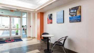 A hotel lobby with a reception desk, a chair, and a table. There is a glass door entrance and a blue exit sign.