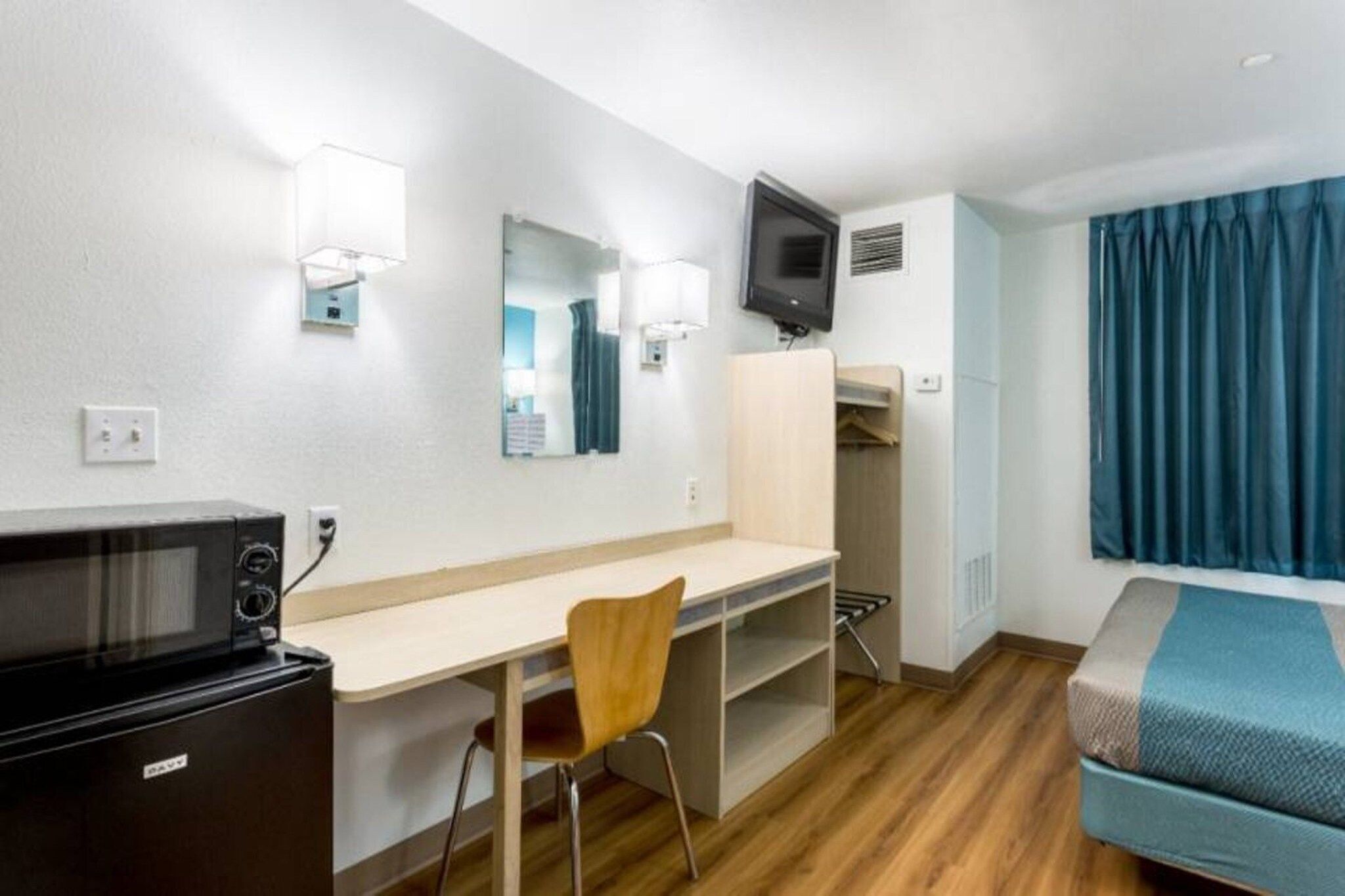 A hotel room with a desk, chair, microwave, TV, wardrobe, and a bed with a blue blanket.