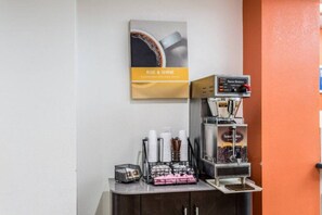 Une machine à café avec une pancarte « RÉVEILLEZ-VOUS ET PASSEZ UNE BONNE JOURNÉE » au-dessus, une pile de gobelets jetables et un panier en métal contenant des ustensiles.