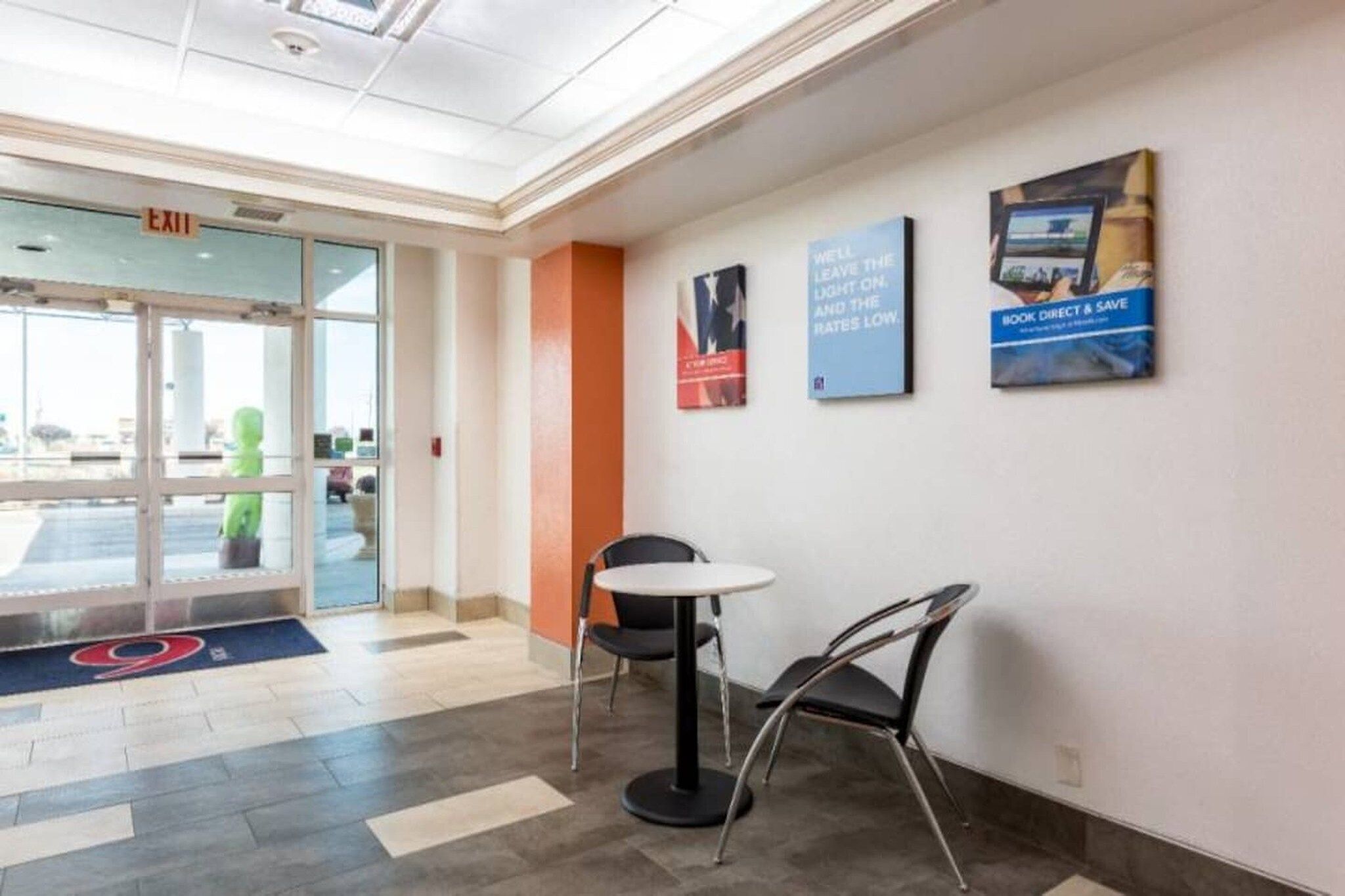 A hotel lobby with a reception desk, a small table, and chairs. There are exit signs and a glass door leading outside.