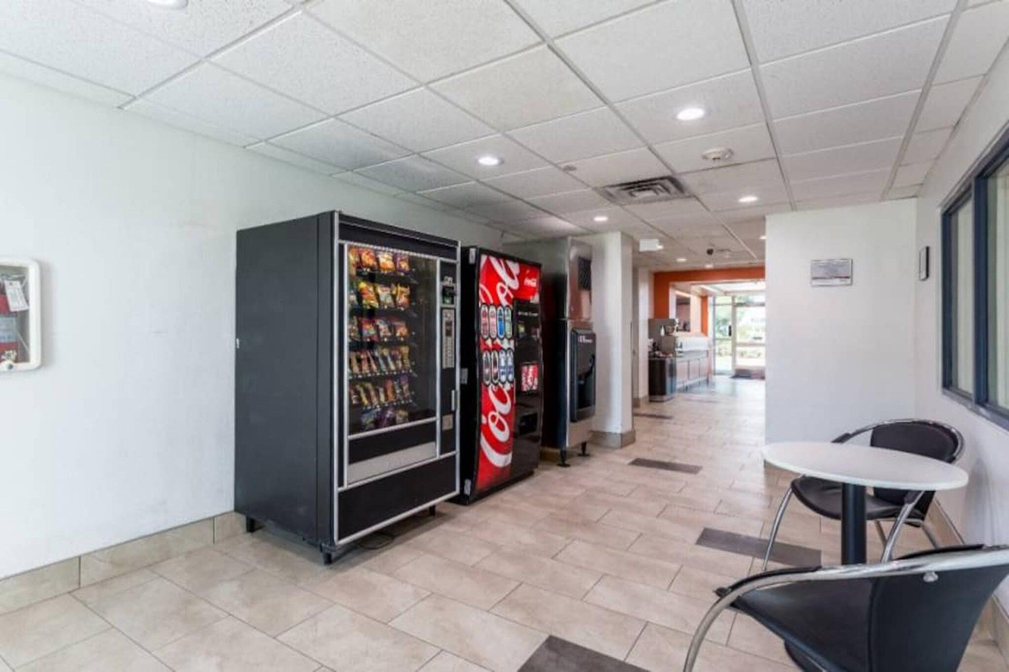 A corridor with vending machines, a seating area with a table and chairs, and a tiled floor.