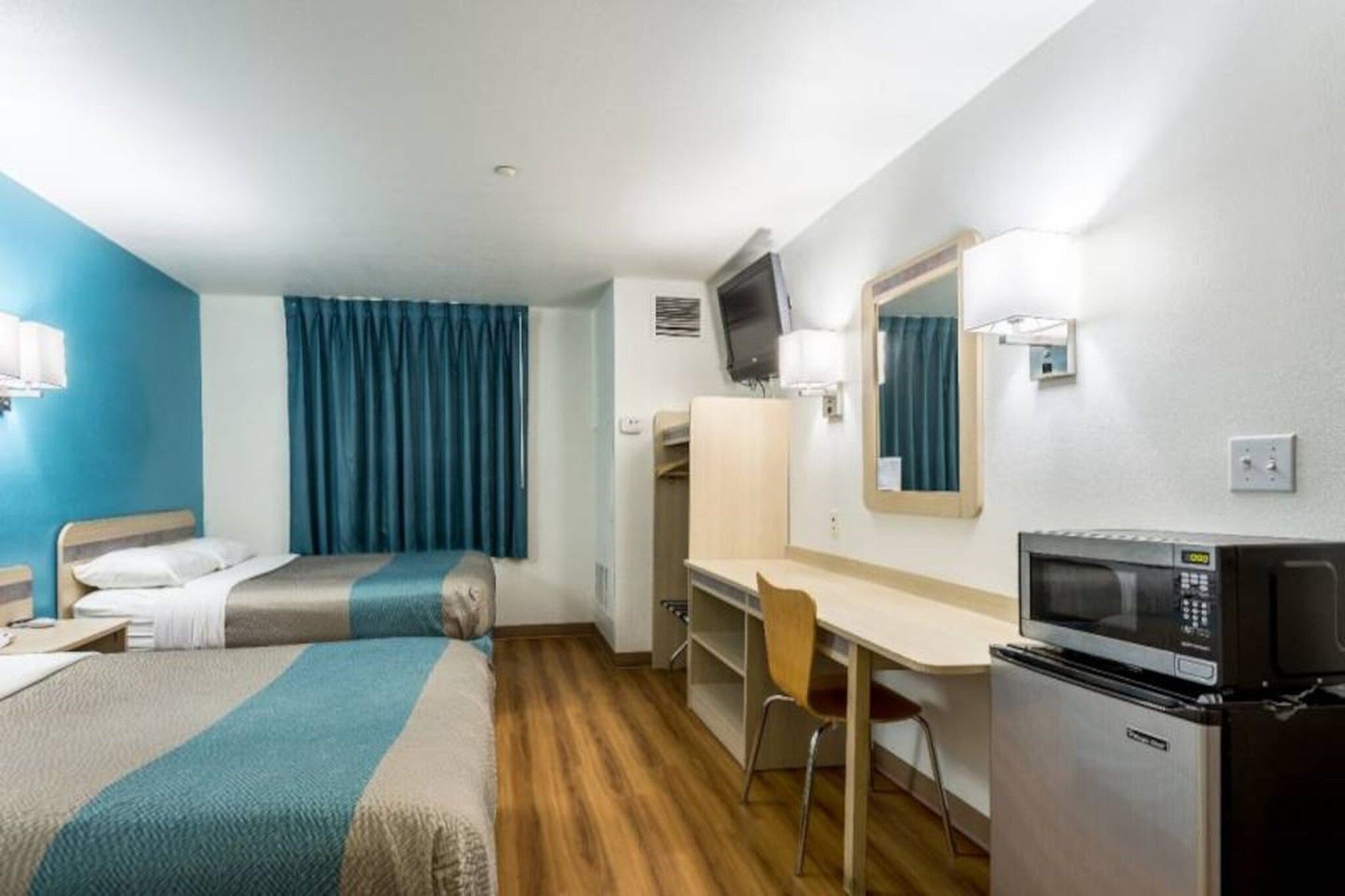 A hotel room with two beds, a desk, a chair, a microwave, and a television.