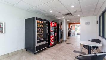 A corridor with vending machines, a seating area with a table and chairs, and a tiled floor.