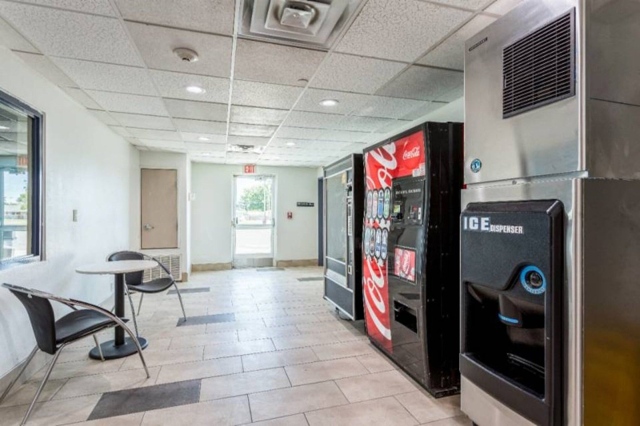 A hotel lobby with a vending machine, a Coca-Cola refrigerator, a table with chairs, and a door in the background.