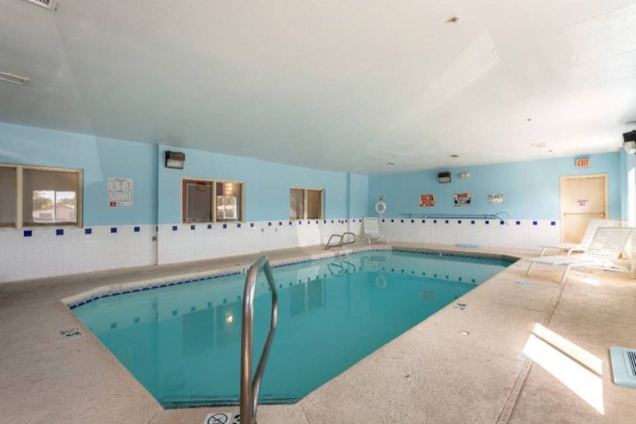 An indoor swimming pool with a tiled floor, a ladder, and a poolside area with chairs.