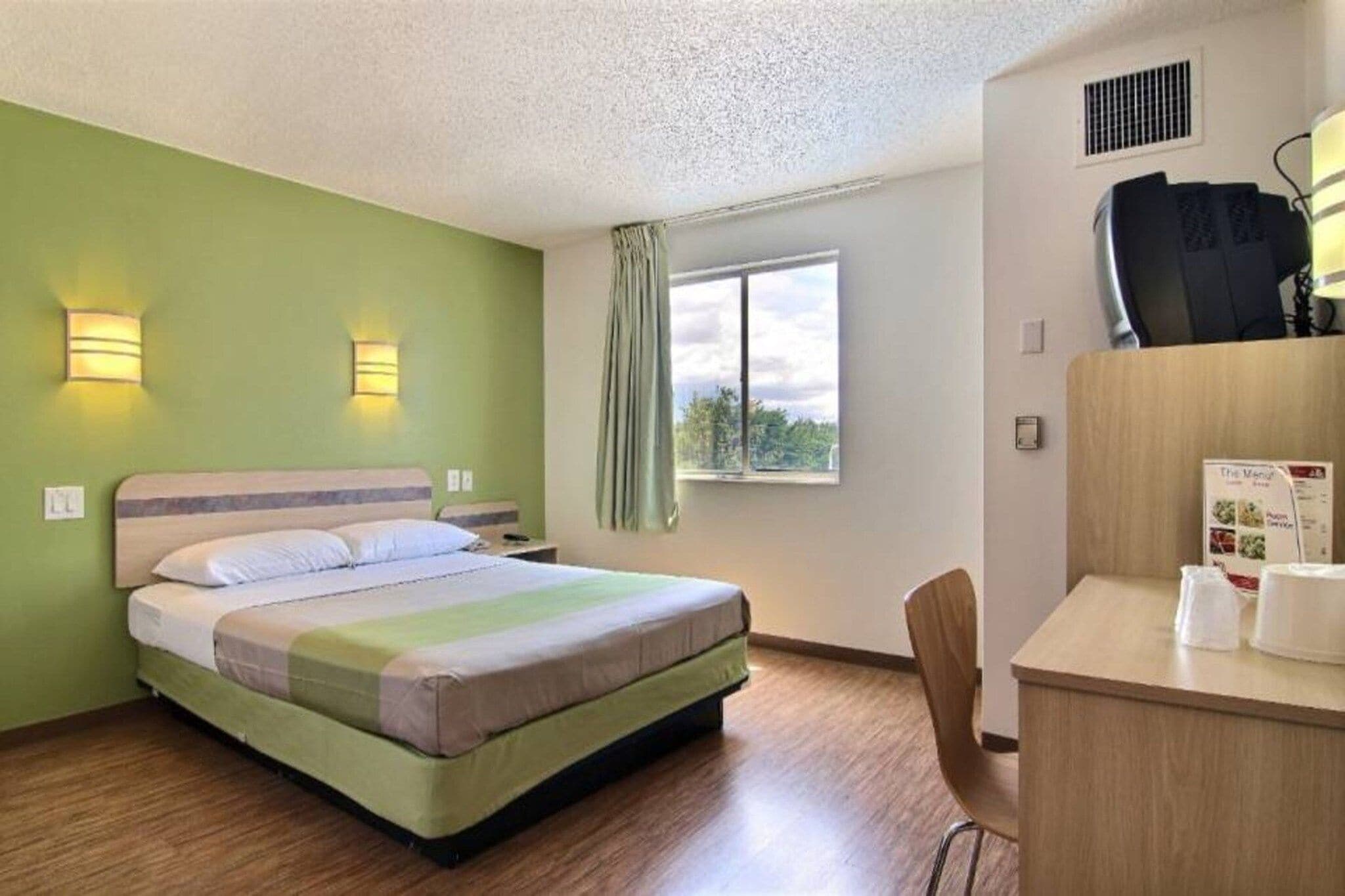 A hotel room with a bed, a desk with a chair, a window with a view, and a wall-mounted air conditioning unit.
