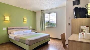 A hotel room with a bed, a desk with a chair, a window with a view, and a wall-mounted air conditioning unit.