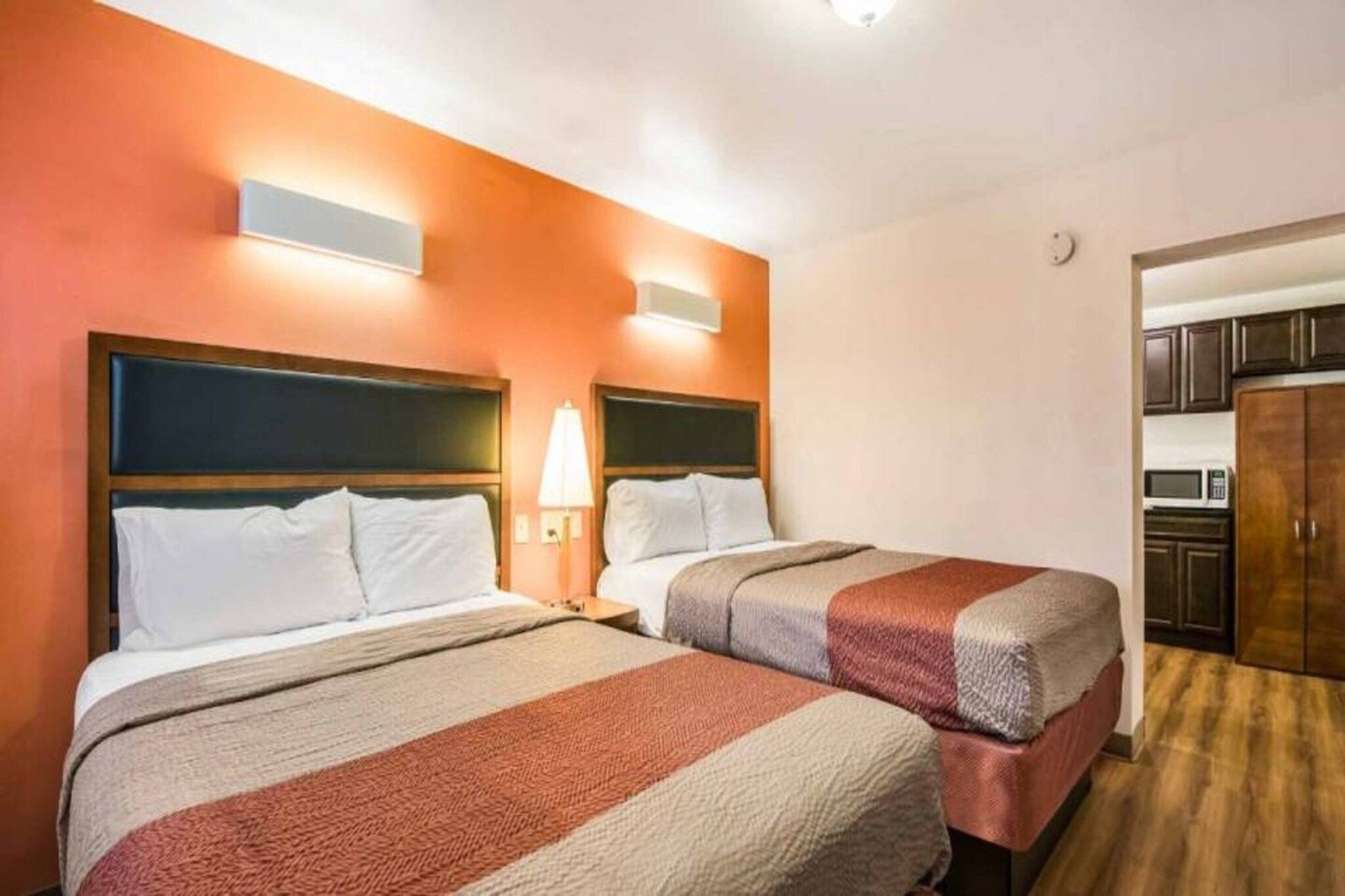 Une chambre d’hôtel avec deux lits, une table de chevet avec une lampe et, au fond, une petite cuisine.