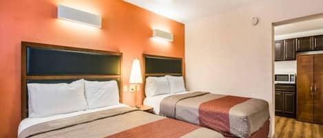 A hotel room with two beds, a nightstand with a lamp, and a kitchenette in the background.